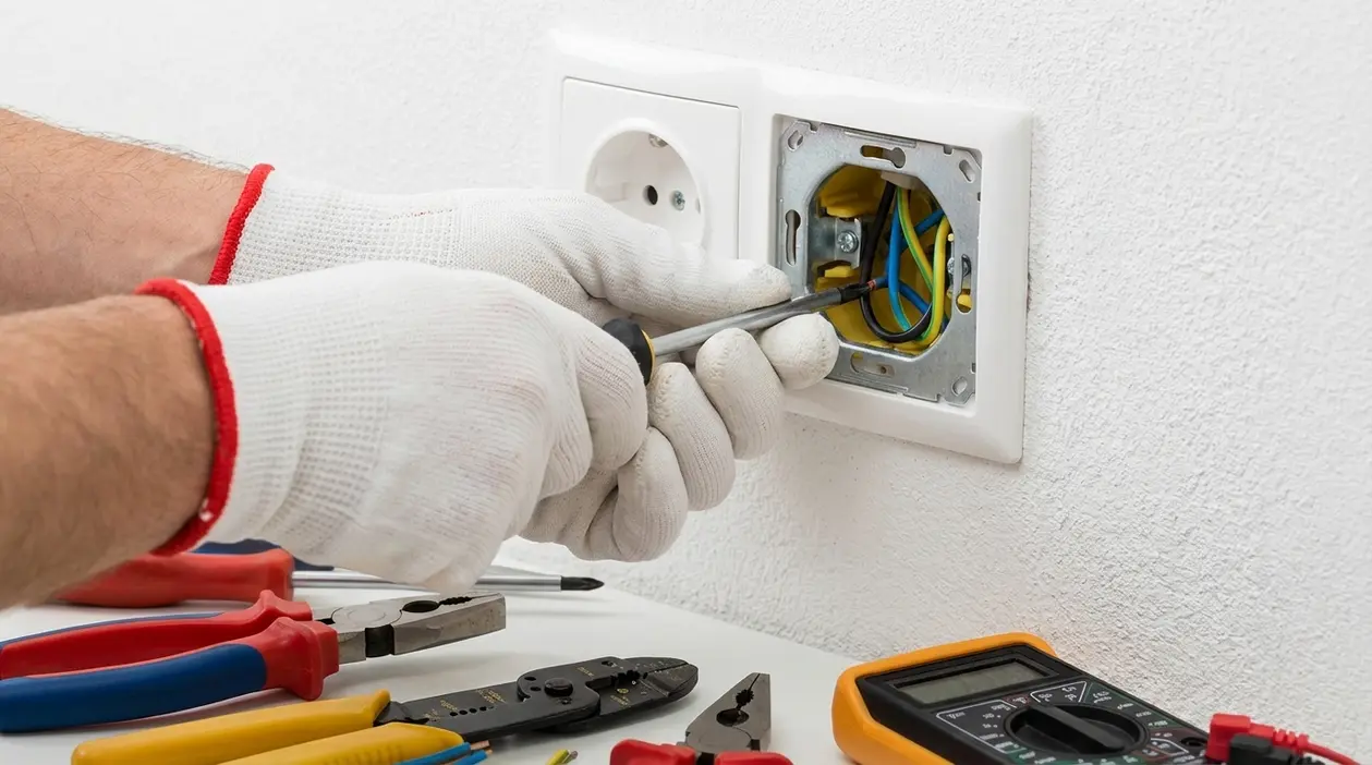 Mani con guanti usano un cacciavite su una presa elettrica aperta per riparare un guasto domestico