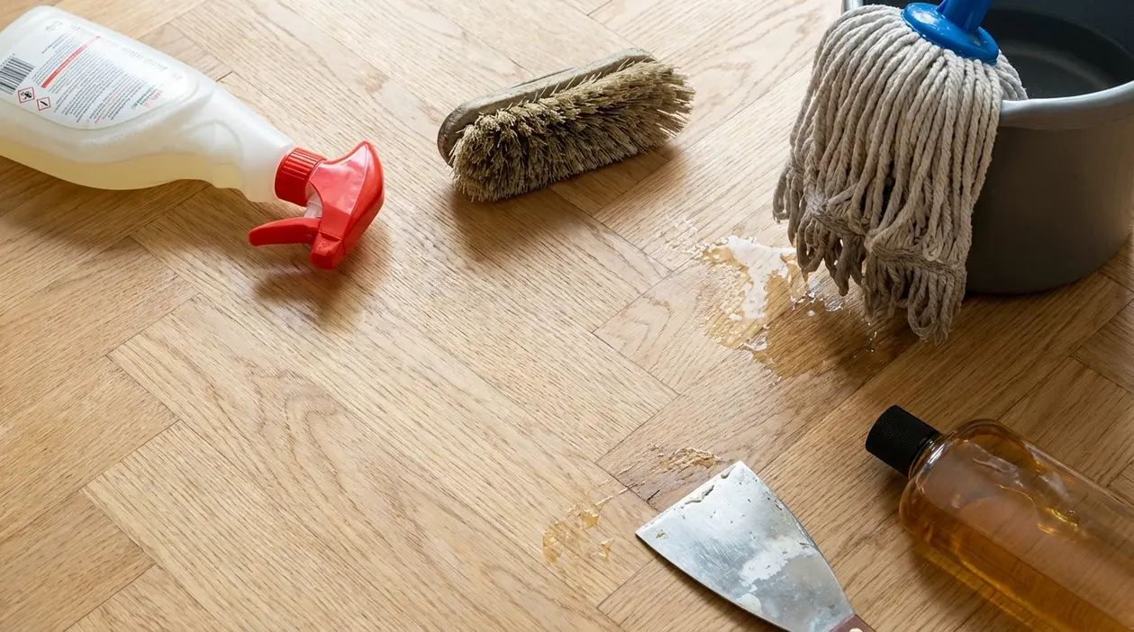 Detergenti, spazzola rigida, mocio bagnato e raschietto appoggiati su un pavimento in parquet