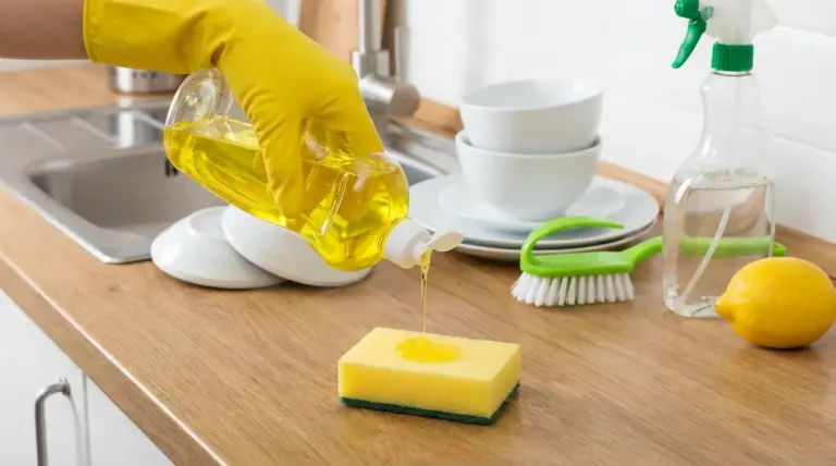 Mano con guanto giallo versa detersivo per piatti su una spugna sul piano cucina vicino al lavello