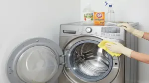 Lavatrice a carica frontale aperta mentre una persona pulisce l’oblò con un panno; prodotti per pulizia sopra